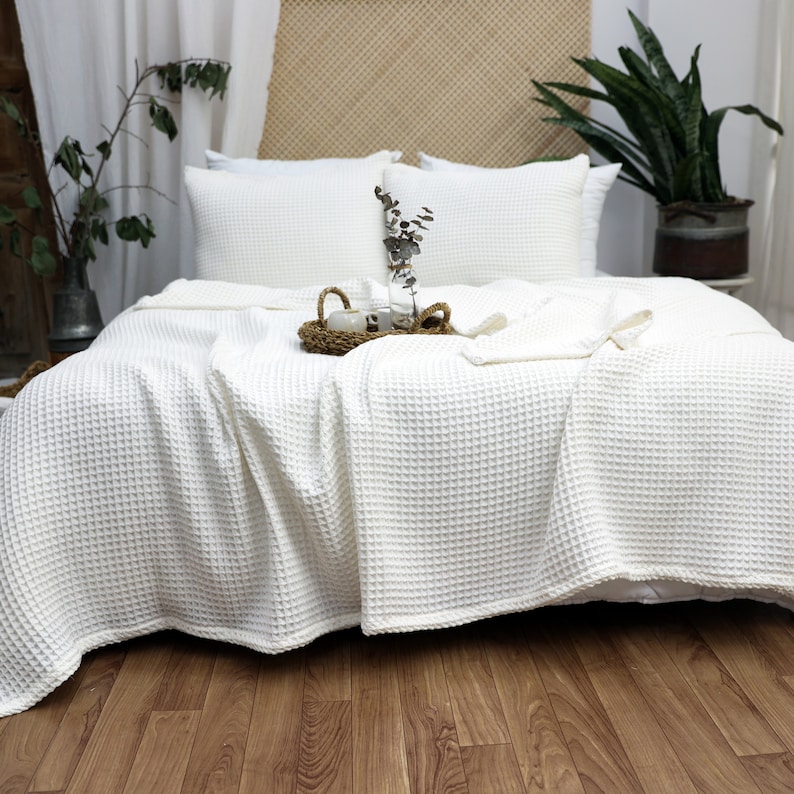 May include: A bed with white waffle-weave bedding, including a blanket and pillows. A small tray with a cup and vase sits on top of the blanket. The bed is in a well-lit room with a wooden floor and a plant in the background.