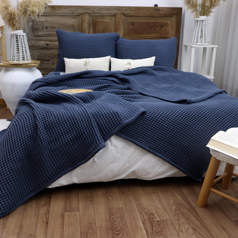 May include: A navy blue waffle-weave blanket and matching pillows are arranged on a bed with white sheets. The bed is against a wooden headboard. Decorative elements include white vases and a small wooden stool.