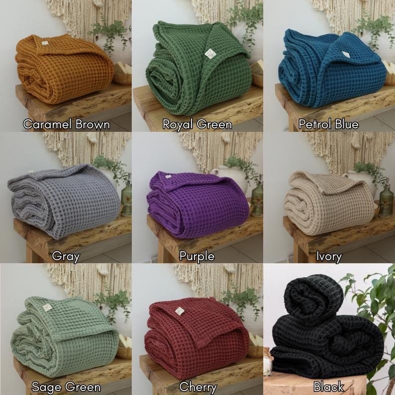 May include: A collection of folded waffle weave blankets in various colors, including caramel brown, royal green, petrol blue, gray, purple, ivory, sage green, cherry, and black. Each blanket is neatly folded and displayed on a wooden surface.
