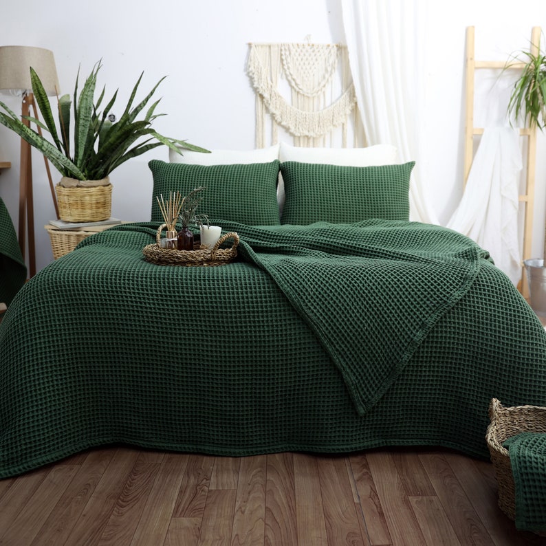 May include: A bed is made with a dark green waffle-weave bedspread and matching pillowcases. A woven tray with candles and decorative items sits on the bed. The room has a natural, earthy aesthetic with plants and woven baskets.