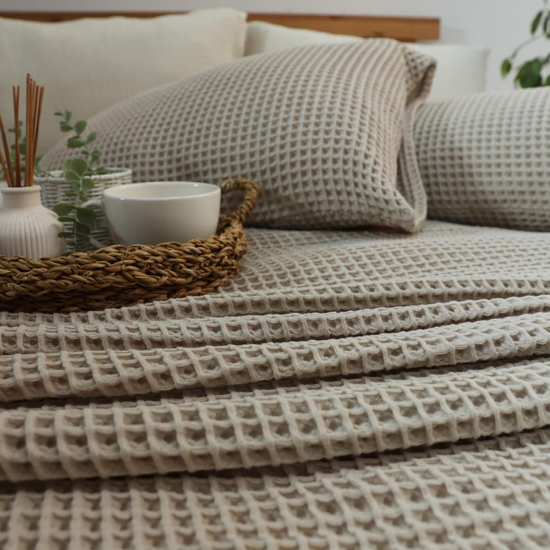 May include: A neutral-toned bedroom scene featuring a bed with a waffle-weave blanket and matching pillows. A woven tray holds a white bowl, a small white vase with reeds, and a small basket with greenery. The bed is made up with neutral-colored linens.