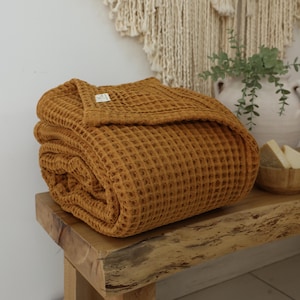 Puede incluir: Una manta de tejido de waffle de color amarillo mostaza doblada en un banco de madera. La manta está hecha de un tejido suave y texturizado.