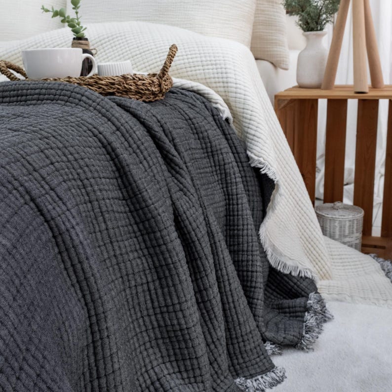May include: A bed with a dark grey textured blanket draped over it, with a white blanket underneath. A woven tray with two white mugs sits on top of the grey blanket. The bed is in a well-lit room.