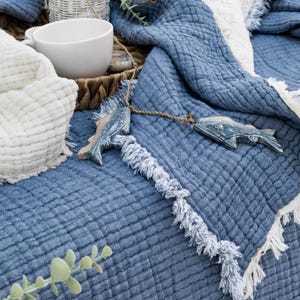 May include: A blue quilted blanket with white fringing is displayed. Two wooden fish ornaments hang from a rope. A white mug and a white folded blanket are in the background. The scene has a coastal, nautical theme.