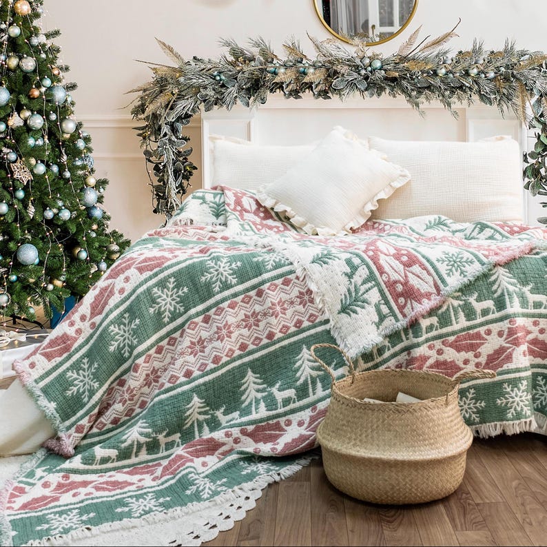 May include: A festive blanket in green, white, and red, featuring a pattern of snowflakes, trees, and reindeer. The blanket is on a bed with white pillows, and a woven basket sits nearby. A Christmas tree is in the background.