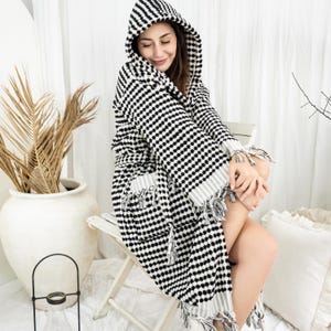 Puede incluir: Una bata con capucha a lunares en blanco y negro. La bata tiene cinturón, bolsillos y flecos. La persona está sentada en una silla blanca. La bata está hecha de un material suave y cómodo.