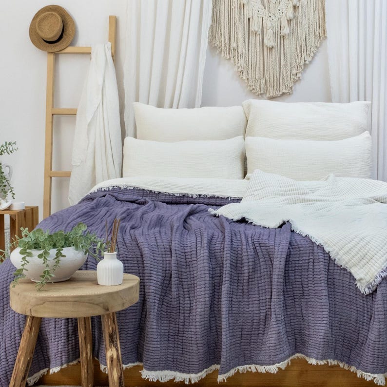 May include: A bed dressed with a lavender textured blanket and white pillows. A white fringed throw blanket is folded on top. A small wooden stool holds a white vase and a bowl of greenery. A straw hat hangs on a wooden ladder.
