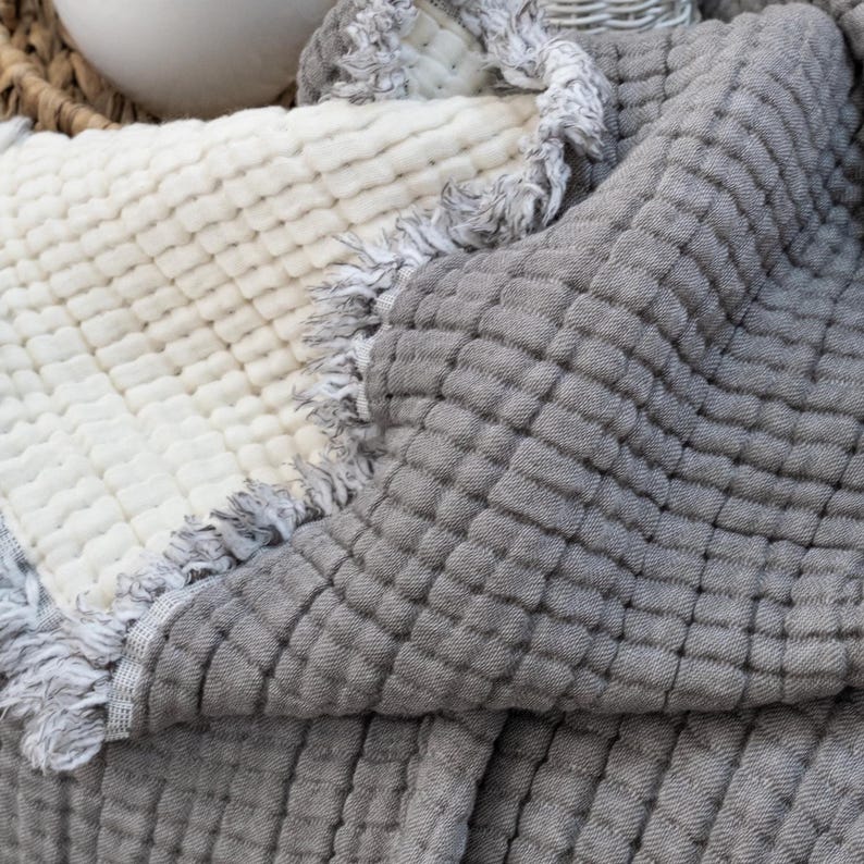 May include: A folded blanket in light grey and off-white. The blanket has a quilted pattern and a fringed edge. The off-white section is on the left, while the grey section is on the right. The blanket is made of a soft material, perfect for home decor.