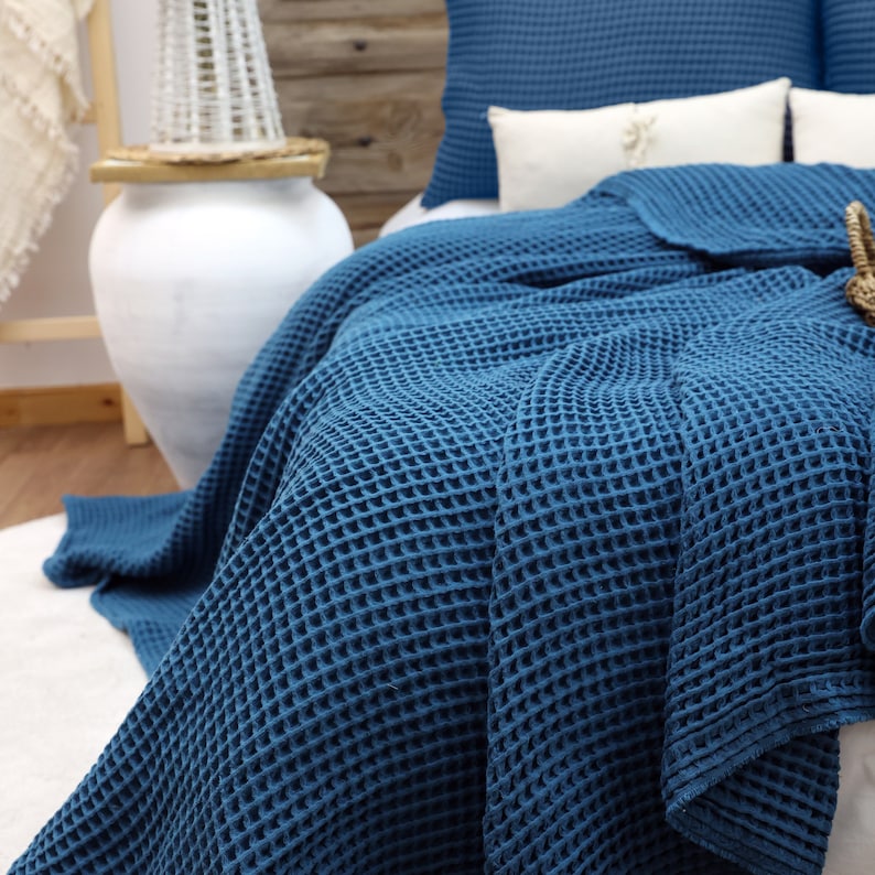 May include: A navy blue waffle-weave blanket draped over a bed, with matching pillows. The blanket's texture is prominent, and the bed is adorned with white pillows and a decorative basket. The scene evokes a cozy, inviting atmosphere.