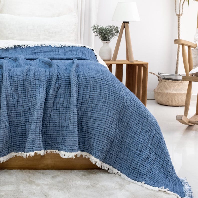 May include: A blue bedspread with a textured, quilted design and a white fringe. The bedspread is draped over a bed with a white pillow. The bed is in a room with a wooden side table and a lamp.