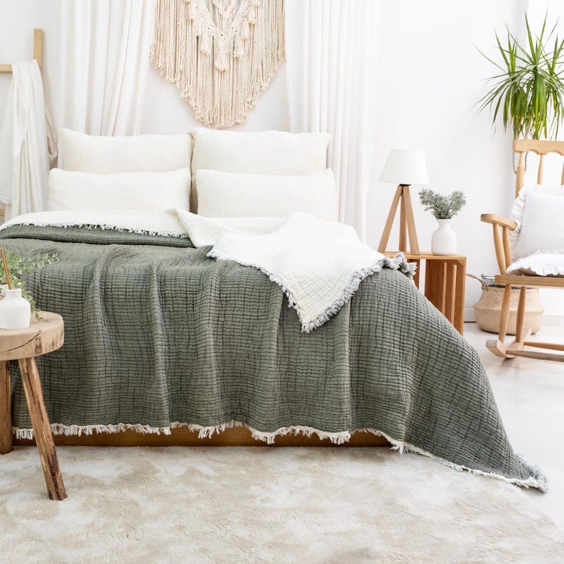May include: A bed is dressed with a sage green blanket with a textured weave and a white fringe. White pillows and a white throw blanket are on the bed. A wooden side table and a rocking chair are in the background.