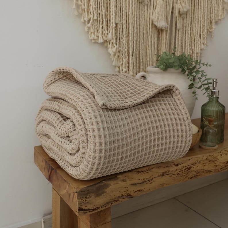 May include: A rolled-up, beige waffle-weave blanket rests on a wooden bench. The blanket's texture is visible, with a grid-like pattern. The scene includes a decorative wall hanging, a white vase with greenery, and a green glass bottle.