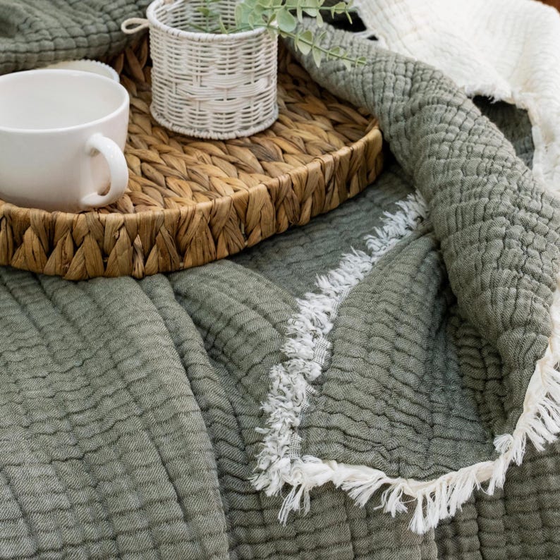 May include: A woven tray holds a white mug, a small white basket with greenery, and rests on a sage green blanket with a white fringe. The blanket is textured and has a quilted appearance, creating a cozy aesthetic.