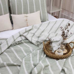 May include: A green and white striped throw blanket with a fringe edge. The blanket is folded on a bed with pillows and a decorative tray with dried flowers.