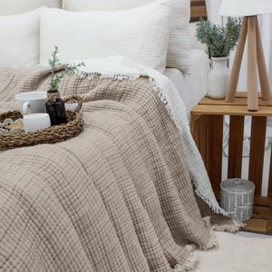 May include: A bed features a beige textured blanket and white pillows. A woven tray holds a cup, a small white container, and a dark bottle. A white fringed blanket is folded over the beige blanket. A wooden side table holds a lamp and a vase.