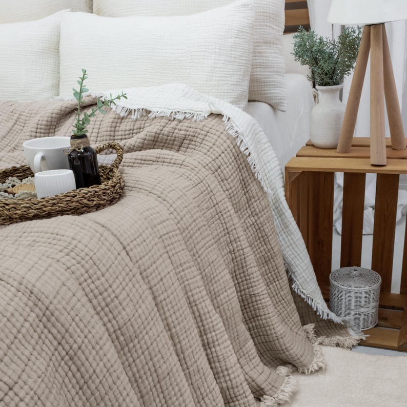 May include: A bed features a beige textured blanket and white pillows. A woven tray holds a cup, a small white container, and a dark bottle. A white fringed blanket is folded over the beige blanket. A wooden side table holds a lamp and a vase.