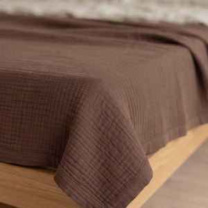 Peut inclure: Une couverture marron foncé avec une texture quadrillée visible drapée sur un cadre de lit en bois clair. La couverture présente une bordure cousue sur les bords et un aspect doux et superposé, recouvrant la surface du lit.