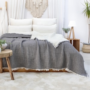 May include: A bed is dressed with a grey blanket with a fringed edge. White pillows and a white fringed blanket are on top. A wooden side table holds a lamp and a vase with greenery. A small wooden stool is in the foreground.