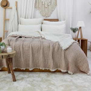 May include: A bed dressed with a beige textured blanket and white pillows. A white fringed throw blanket is on top. A wooden side table holds a lamp and a vase with greenery. A wooden ladder and macrame wall hanging are in the background.