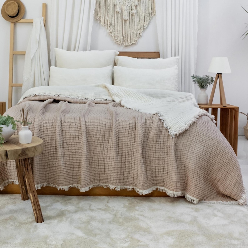 May include: A bed dressed with a beige textured blanket and white pillows. A white fringed throw blanket is on top. A wooden side table holds a lamp and a vase with greenery. A wooden ladder and macrame wall hanging are in the background.