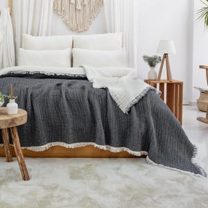 May include: A bed is dressed with a dark grey textured blanket with a white fringe. White pillows and a white blanket are on the bed. The bed is in a bright room with a white rug and a wooden side table.