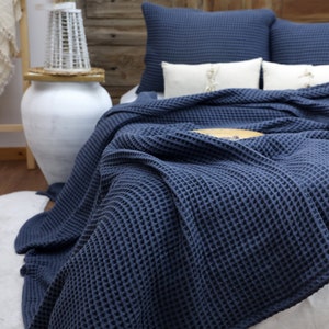 May include: A navy blue waffle-weave blanket draped over a bed, with matching pillows. The blanket's texture is prominent, and the bed is made with white sheets. Decorative pillows with tassels are also visible.
