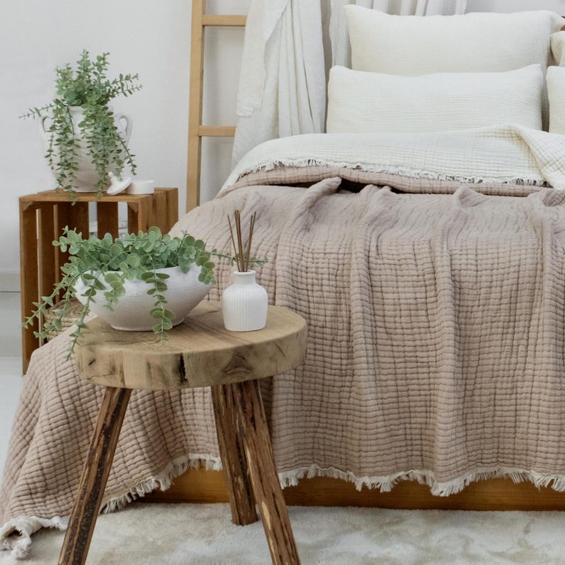 May include: A bedroom setting with a bed covered in a beige textured blanket and white pillows. A small wooden side table supports a white vase and a bowl with greenery. A wooden crate and ladder are also present.