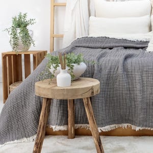 May include: A bedroom scene with a grey textured blanket on a bed. A small wooden stool supports a white vase with reeds and greenery. A wooden crate and a white vase with greenery are also visible.