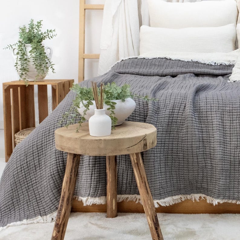 May include: A bedroom scene with a grey textured blanket on a bed. A small wooden stool supports a white vase with reeds and greenery. A wooden crate and a white vase with greenery are also visible.