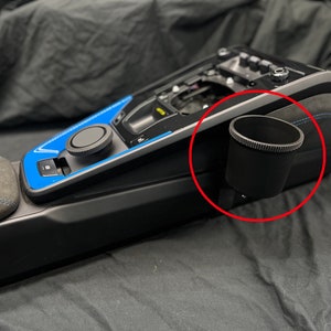 May include: A black cup holder is attached to the center console of a car. The cup holder is made of plastic and has a textured surface. The console is black and has a blue accent.