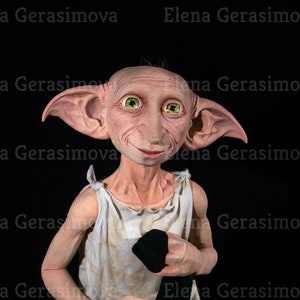 Puede incluir: Una peque&ntilde;a figura de elfo de piel p&aacute;lida, con orejas grandes y ojos verdes, lleva un vestido blanco sin mangas y sostiene un calcet&iacute;n negro. La figura est&aacute; de pie sobre una plataforma de madera.