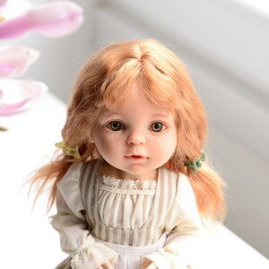 Puede incluir: Una mu&ntilde;eca de porcelana con el pelo largo, ondulado y casta&ntilde;o claro, que lleva un vestido a rayas blancas y marrones con un ribete de encaje. La mu&ntilde;eca tiene ojos verdes y una expresi&oacute;n dulce.