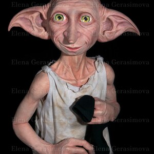 Puede incluir: Un primer plano de una mu&ntilde;eca de Dobby el elfo dom&eacute;stico. La mu&ntilde;eca lleva una camisa blanca sin mangas y calcetines negros. La mu&ntilde;eca tiene orejas grandes, ojos verdes y una sonrisa traviesa.