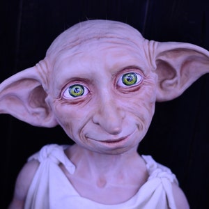 May include: A close-up of a Dobby the House-Elf doll with large pointed ears, a white robe, and green eyes. The doll is made of a soft, realistic material and has a friendly expression.