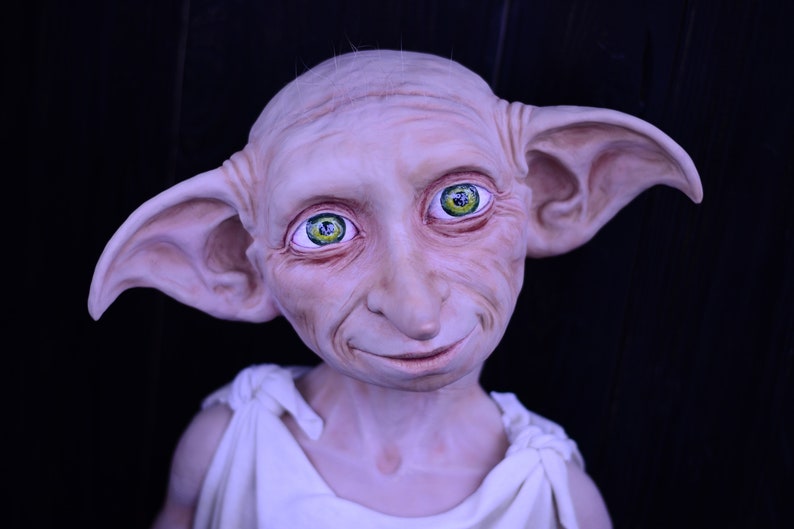 May include: A close-up of a Dobby the House-Elf doll with large pointed ears, a white robe, and green eyes. The doll is made of a soft, realistic material and has a friendly expression.