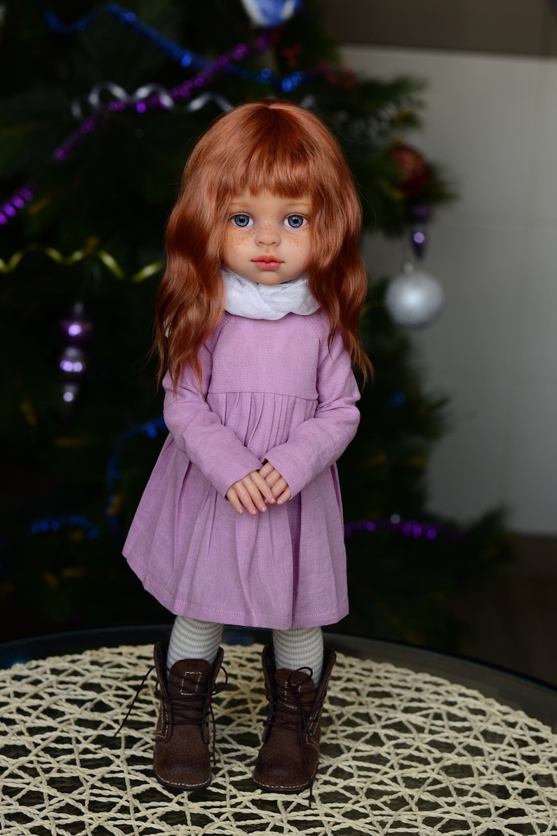 May include: A doll with red hair wearing a lavender dress, white scarf, and brown boots. The doll is standing on a woven mat.