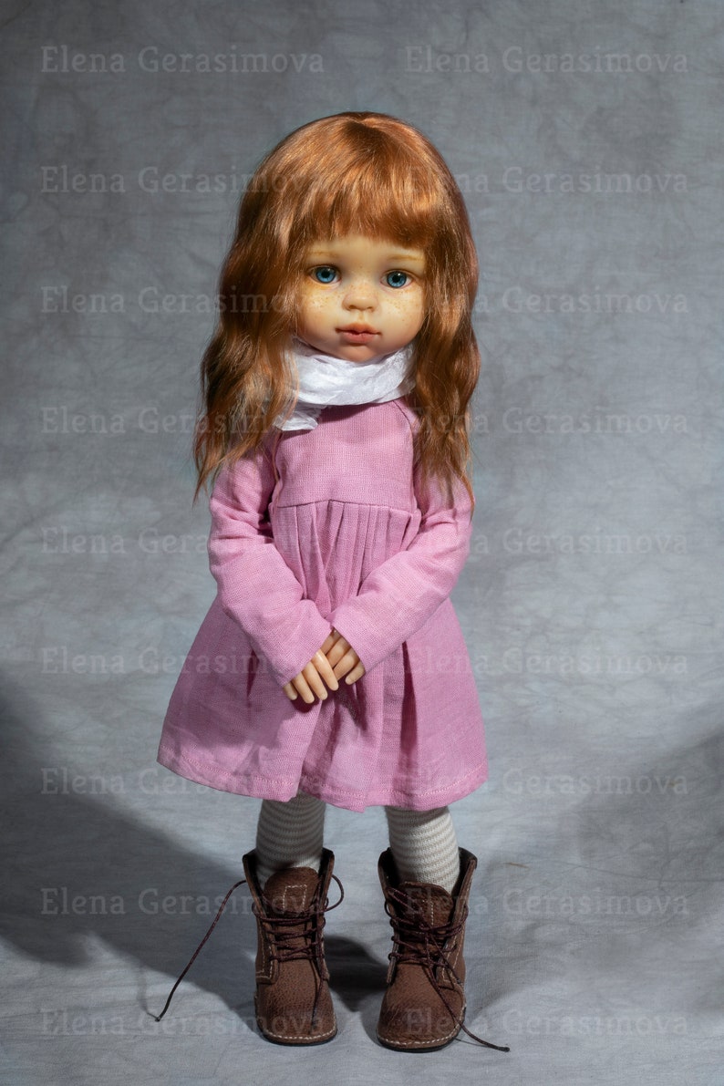 May include: A doll with long, wavy, auburn hair, wearing a pink dress, white scarf, and brown boots. The doll has blue eyes and a freckled face.