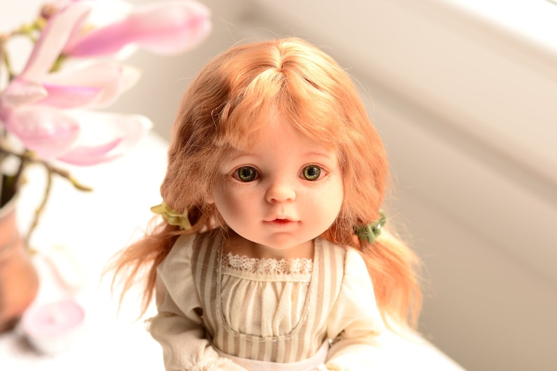 Puede incluir: Una mu&ntilde;eca de porcelana con el pelo largo y rojo, peinado en trenzas. La mu&ntilde;eca lleva un vestido a rayas blancas y marrones con un ribete de encaje. La mu&ntilde;eca tiene ojos verdes y una expresi&oacute;n dulce.