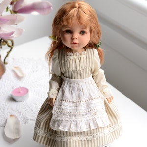 Puede incluir: Una mu&ntilde;eca de porcelana con el pelo rojo que lleva un vestido a rayas con un delantal blanco y zapatos marrones.