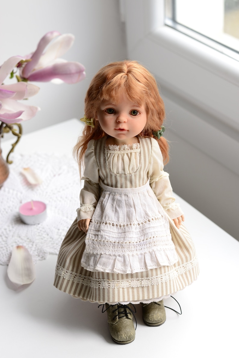 Puede incluir: Una mu&ntilde;eca de porcelana con el pelo rojo que lleva un vestido a rayas con un delantal blanco y zapatos marrones.