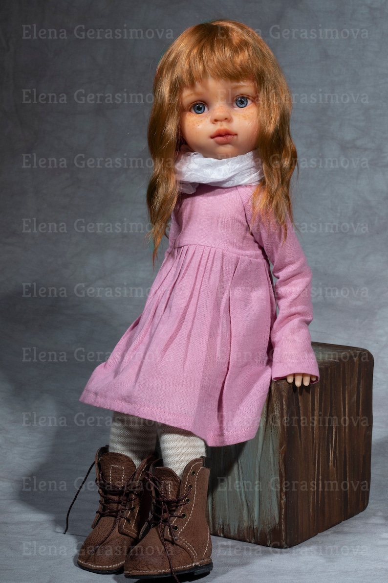May include: A doll with long, wavy, auburn hair, wearing a pink dress, white scarf, and brown boots. The doll is sitting on a wooden box.