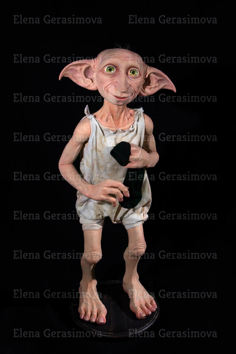 Puede incluir: Una peque&ntilde;a figura de elfo de piel p&aacute;lida, con orejas grandes y ojos verdes, lleva un vestido blanco sin mangas y sostiene un calcet&iacute;n negro. La figura est&aacute; de pie sobre una plataforma de madera.