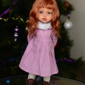 May include: A doll with long, wavy red hair, wearing a pink dress, white scarf, and brown boots. The doll is standing in front of a Christmas tree.