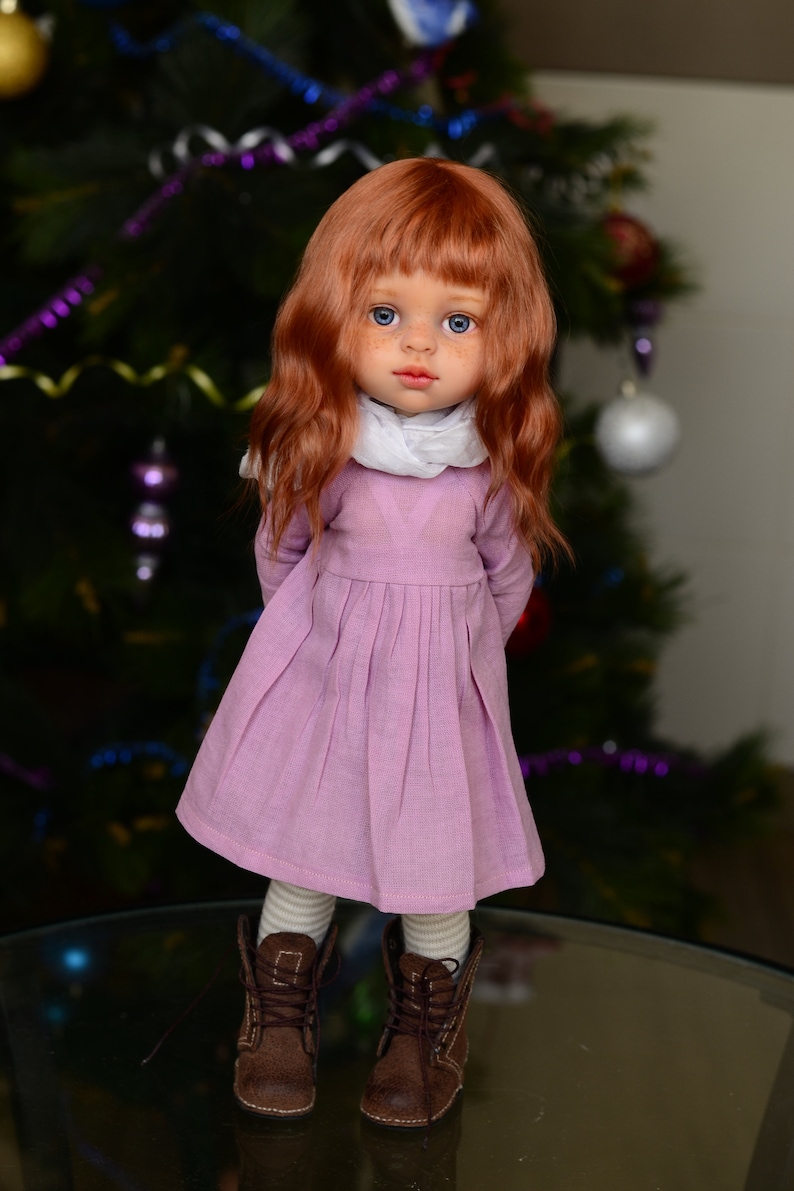 May include: A doll with long, wavy red hair, wearing a pink dress, white scarf, and brown boots. The doll is standing in front of a Christmas tree.