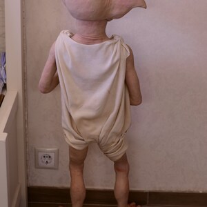 Puede incluir: Una mu&ntilde;eca de elfo de piel clara con una t&uacute;nica blanca de pie en un suelo de madera. La mu&ntilde;eca tiene orejas grandes y est&aacute; de espaldas a la c&aacute;mara.