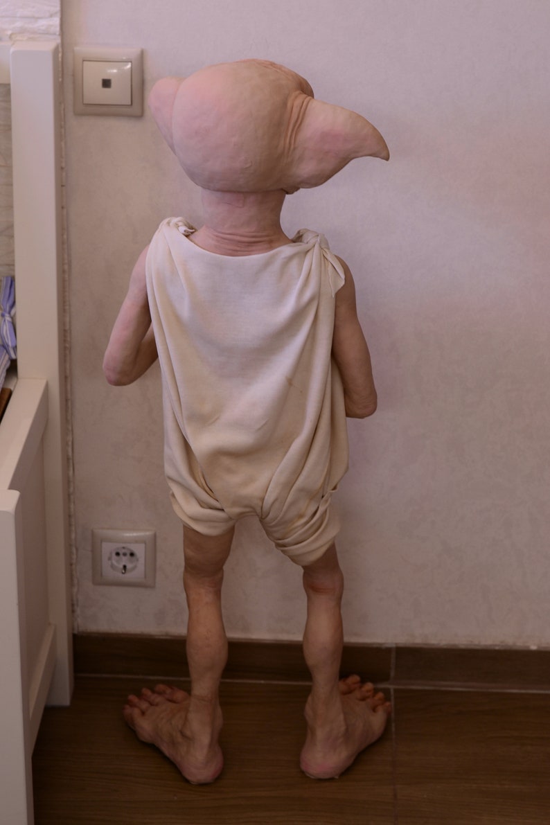 Puede incluir: Una mu&ntilde;eca de elfo de piel clara con una t&uacute;nica blanca de pie en un suelo de madera. La mu&ntilde;eca tiene orejas grandes y est&aacute; de espaldas a la c&aacute;mara.