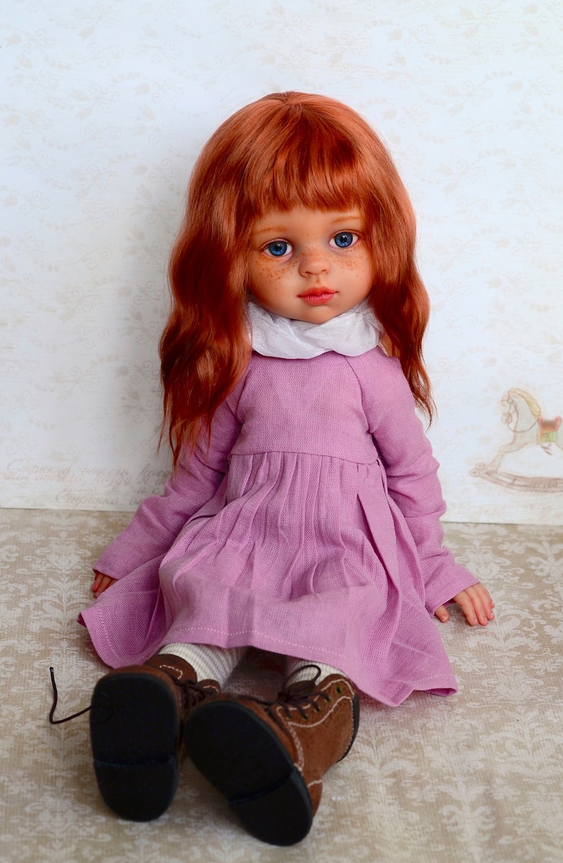 May include: A red-haired doll wearing a pink dress and brown shoes. The doll has blue eyes and freckles.