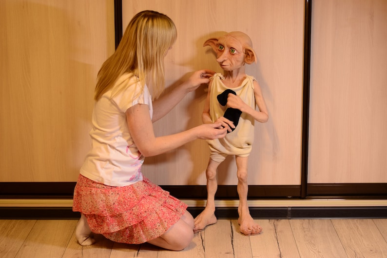 Puede incluir: Una mujer con un vestido rosa y blanco est&aacute; arrodillada y ayudando a una mu&ntilde;eca vestida con una t&uacute;nica blanca y calcetines negros. La mu&ntilde;eca tiene orejas grandes y una cara puntiaguda.