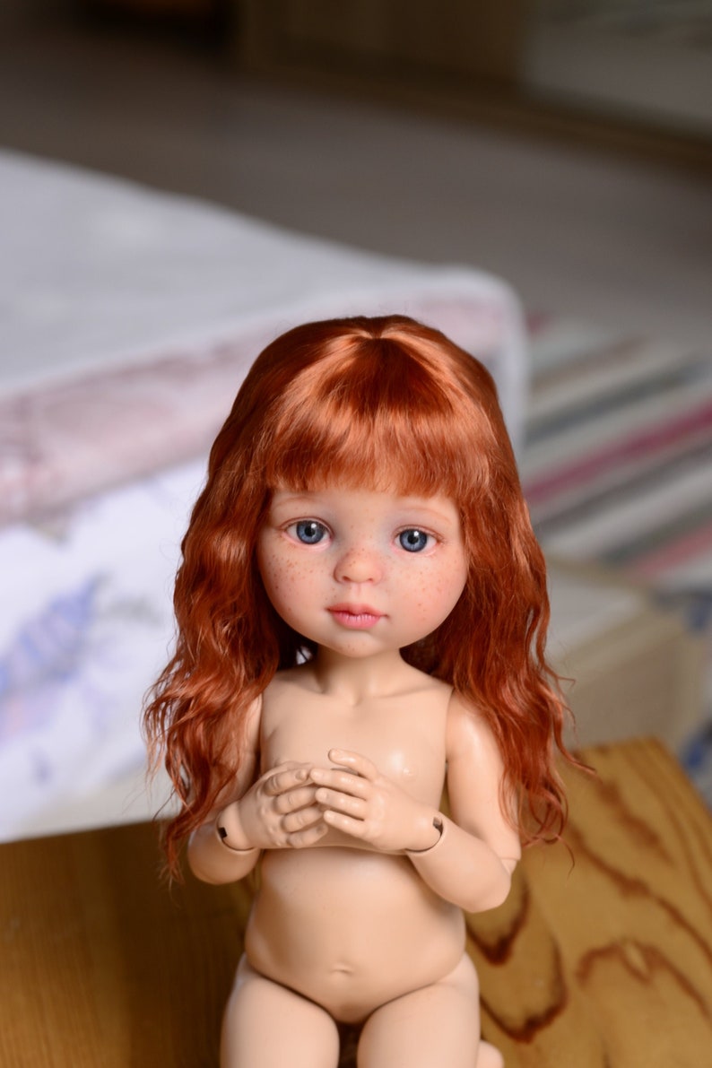 May include: A doll with long, wavy, red hair and blue eyes. The doll is wearing a nude-colored outfit and has freckles on its face.