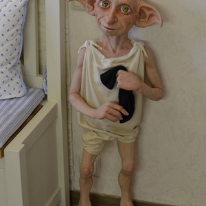 May include: A whimsical elf doll with large pointed ears, green eyes, and a light brown complexion. The doll is wearing a white tunic with a black sock tucked into the front. The doll is standing on a wooden floor.